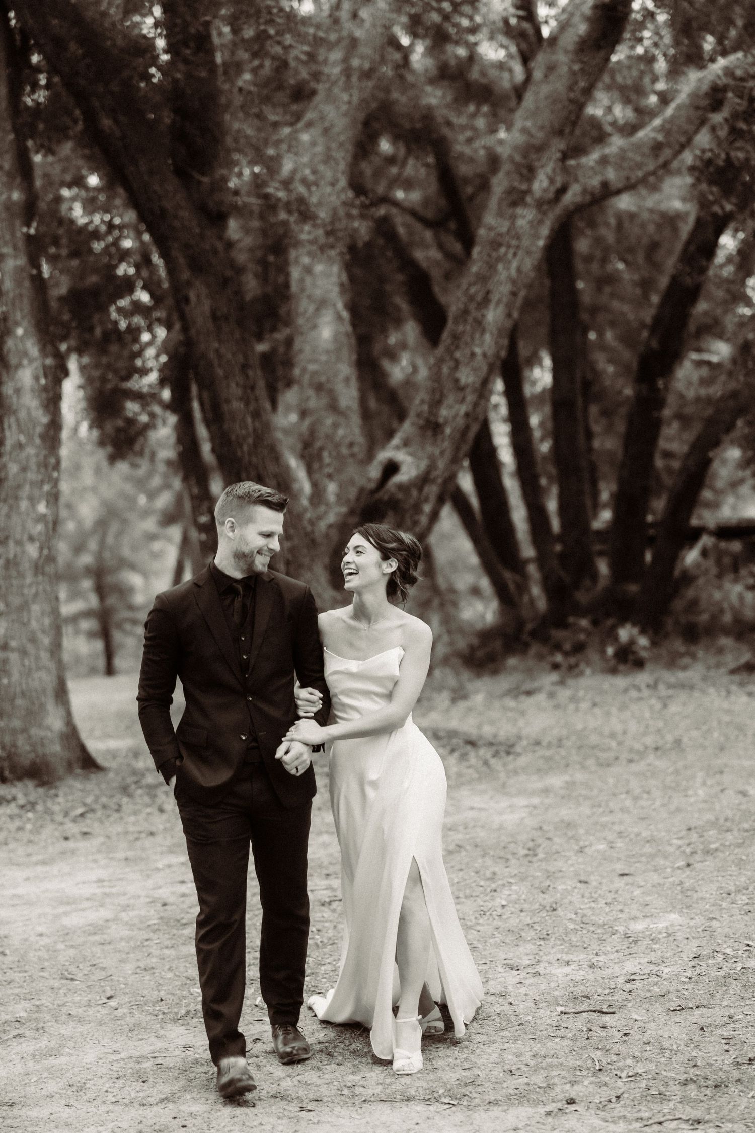 Black and white photo of a wedding couple walking and laughing together on a tree-lined path.