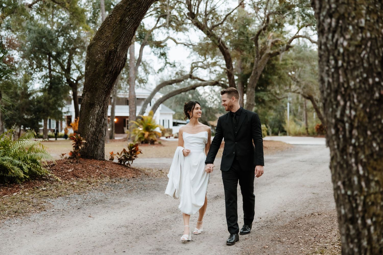 Couple walks along tree-lined path at rustic outdoor venue.