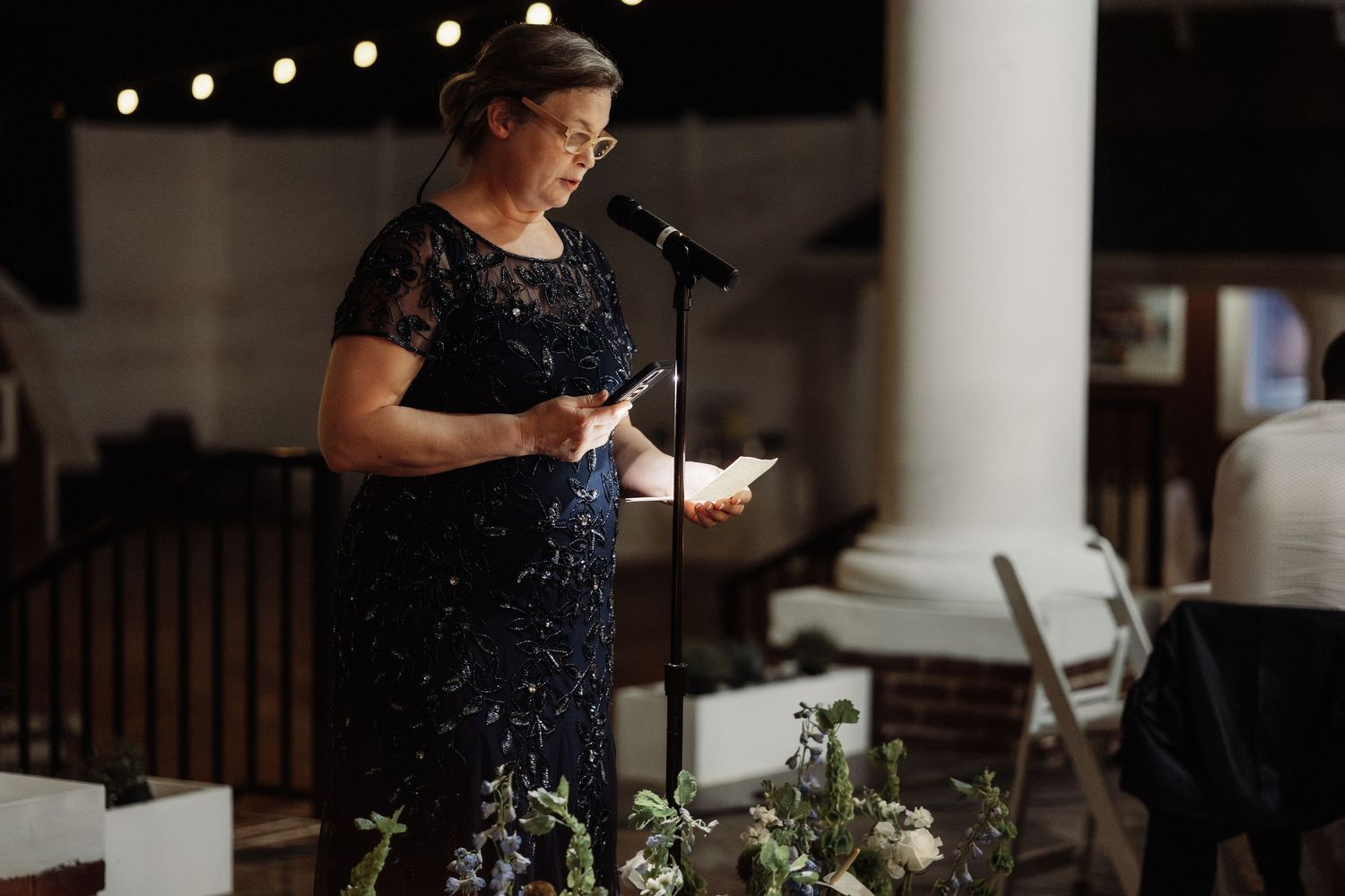 A guest speaker reads from notes at an evening wedding reception with string lights in the background.