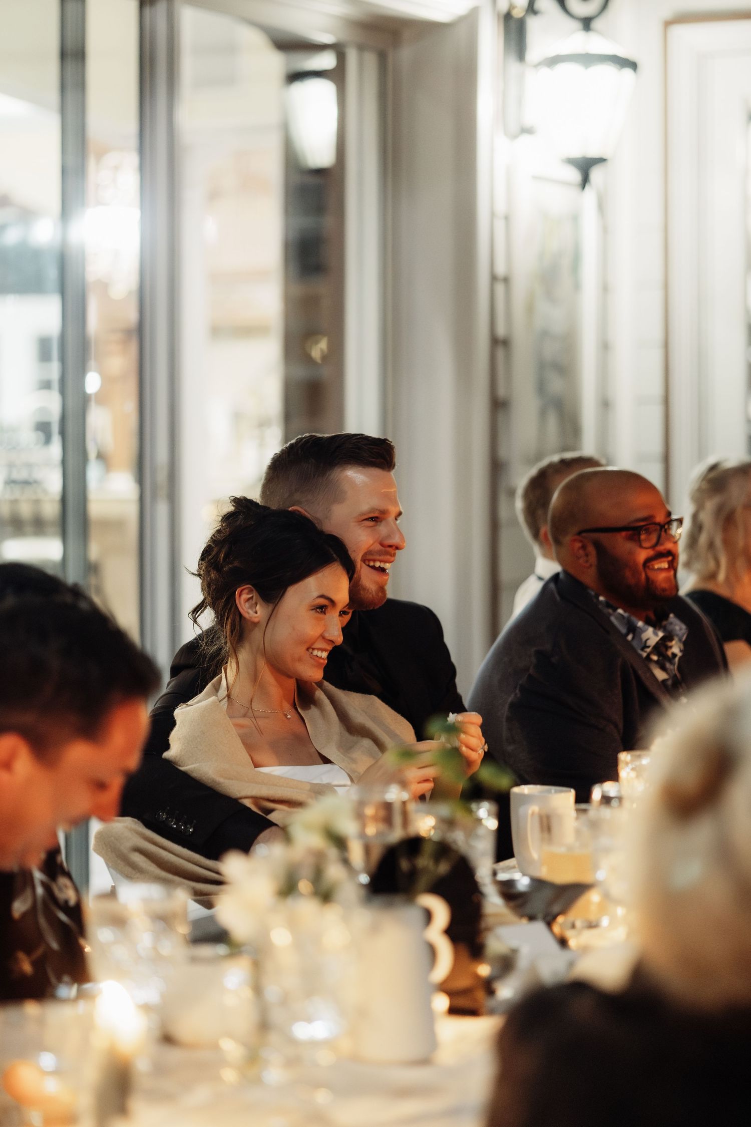 People gathered around a candlelit dinner table sharing joyful conversation at an indoor evening event.