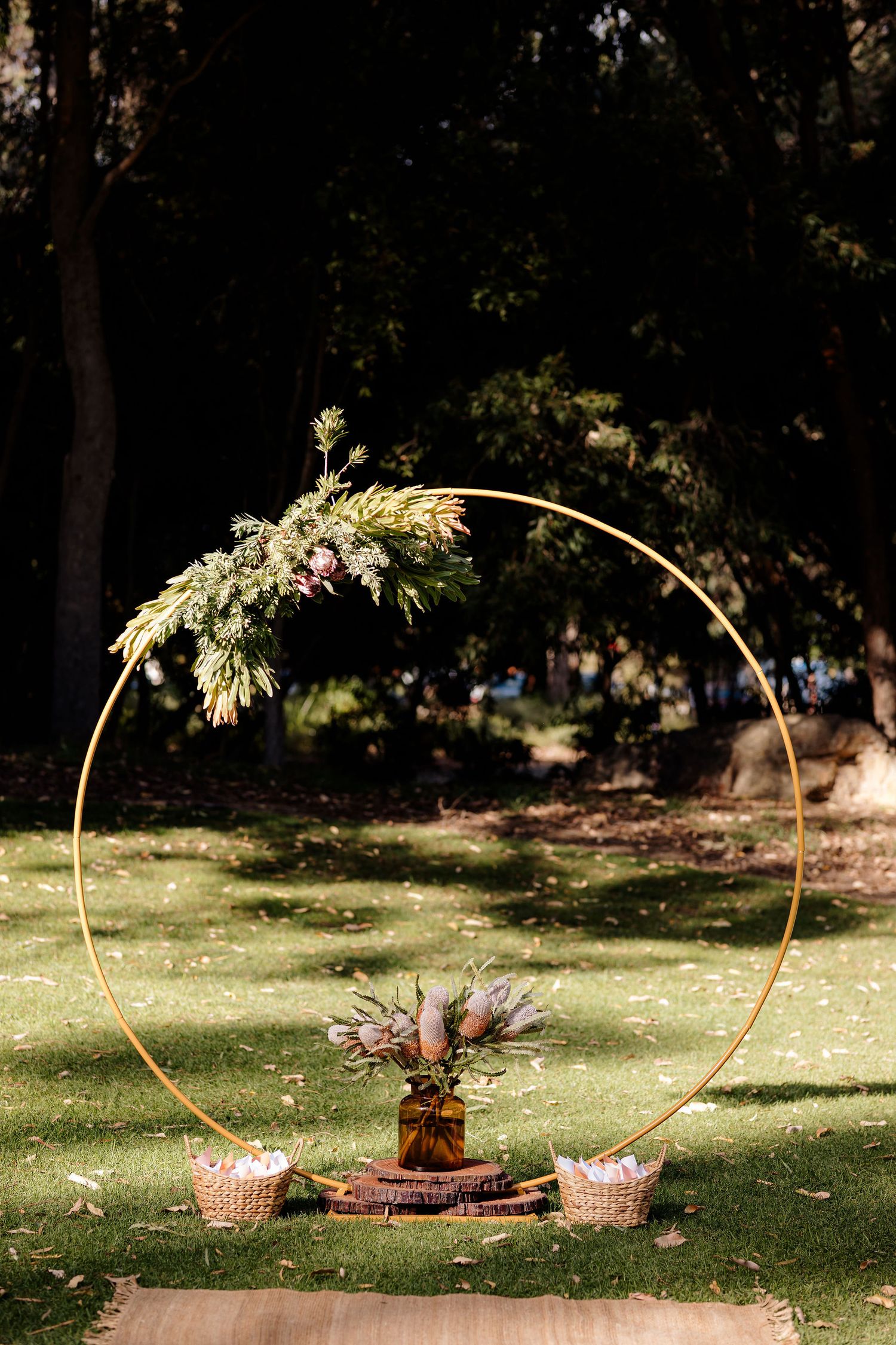 A circular gold wedding arch with floral arrangements stands on grass with decorative baskets and picnic setup below.
