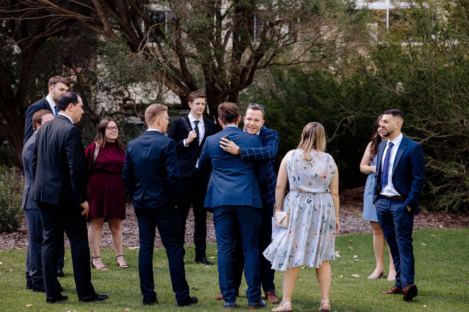 Multiple wedding guests in formal attire stand together in conversation outside on a green lawn during a daytime event.