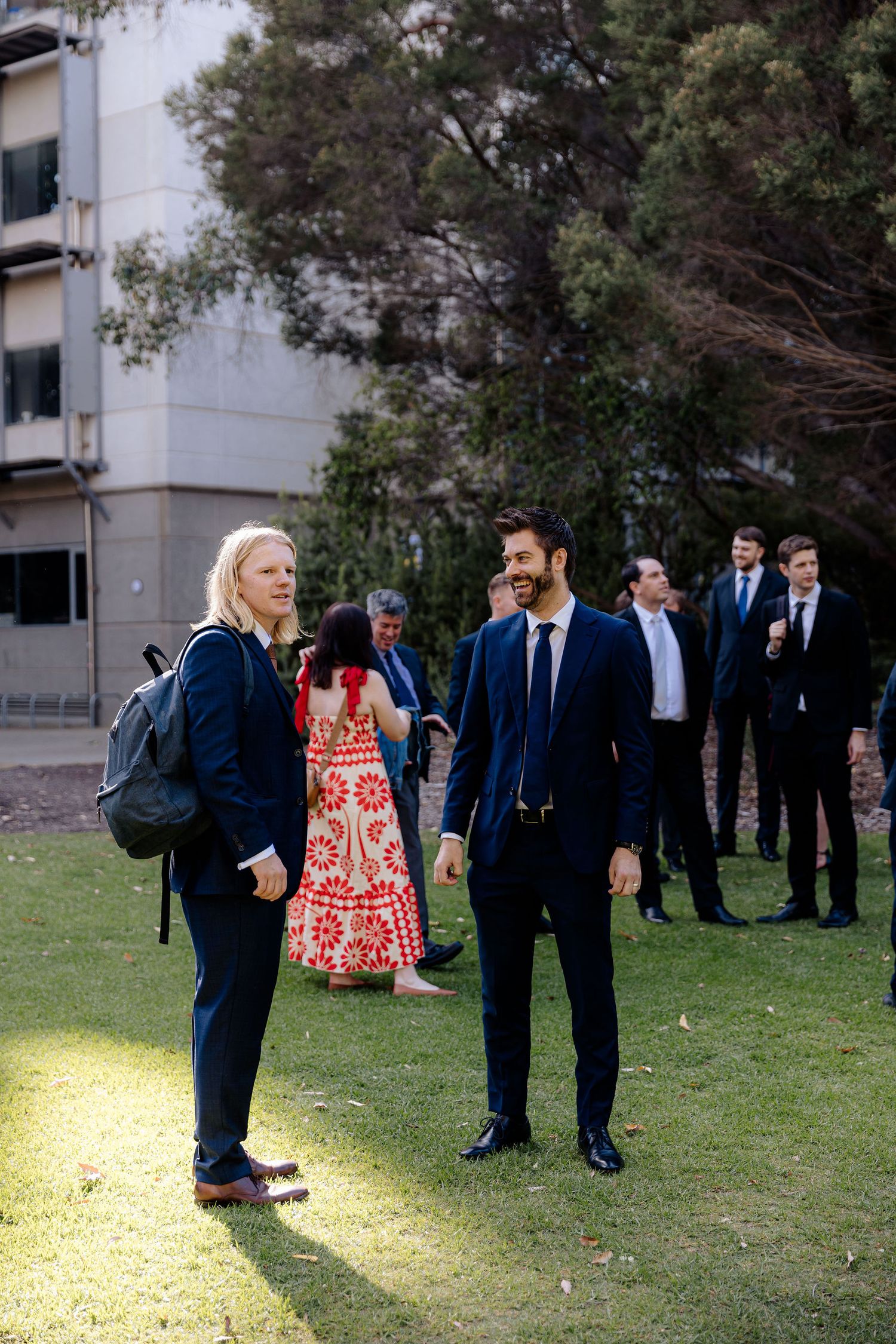 People in formal attire stand on a grassy lawn during what appears to be an outdoor gathering.