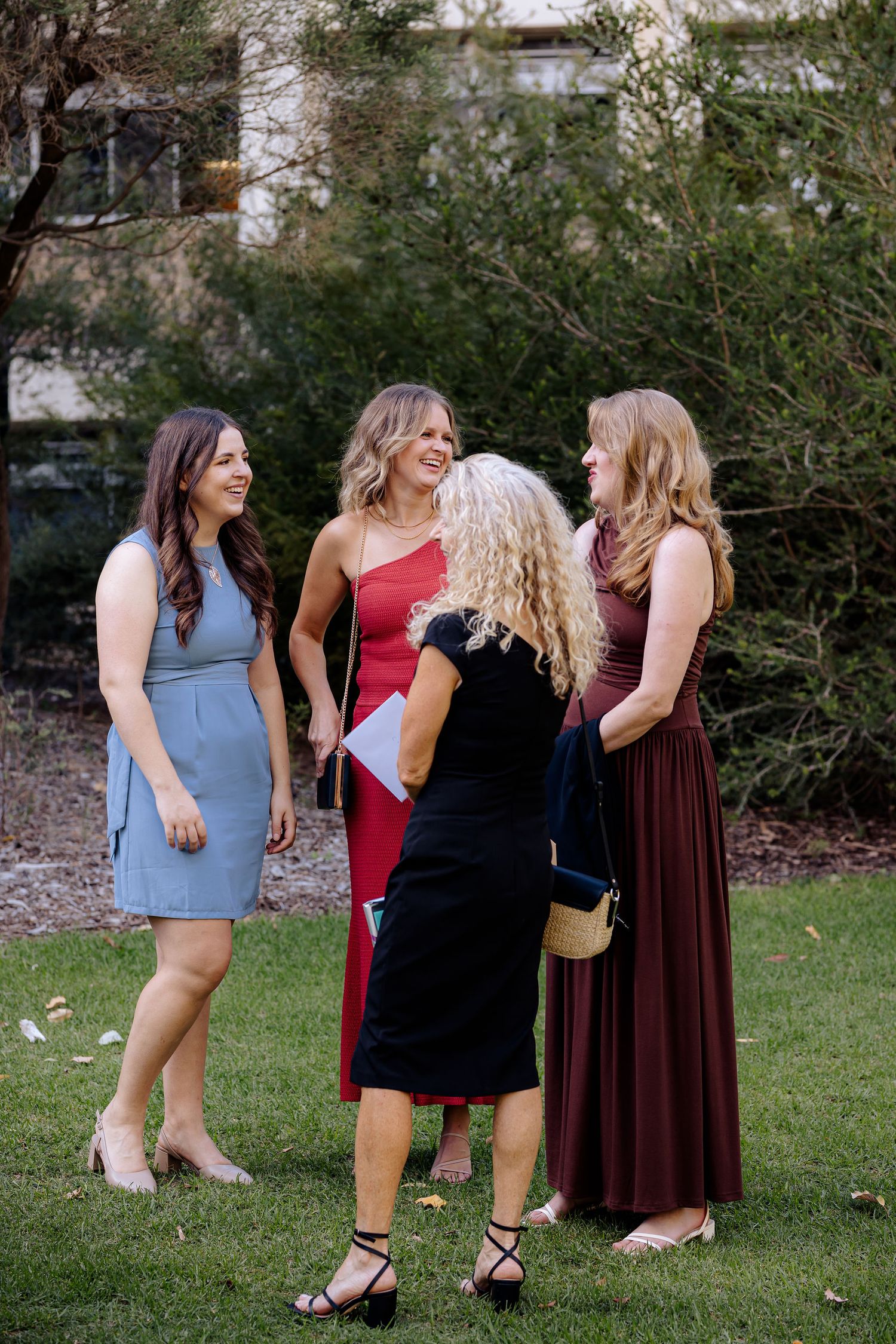 A group of women in formal dresses stand chatting together on a lawn with greenery in the background.