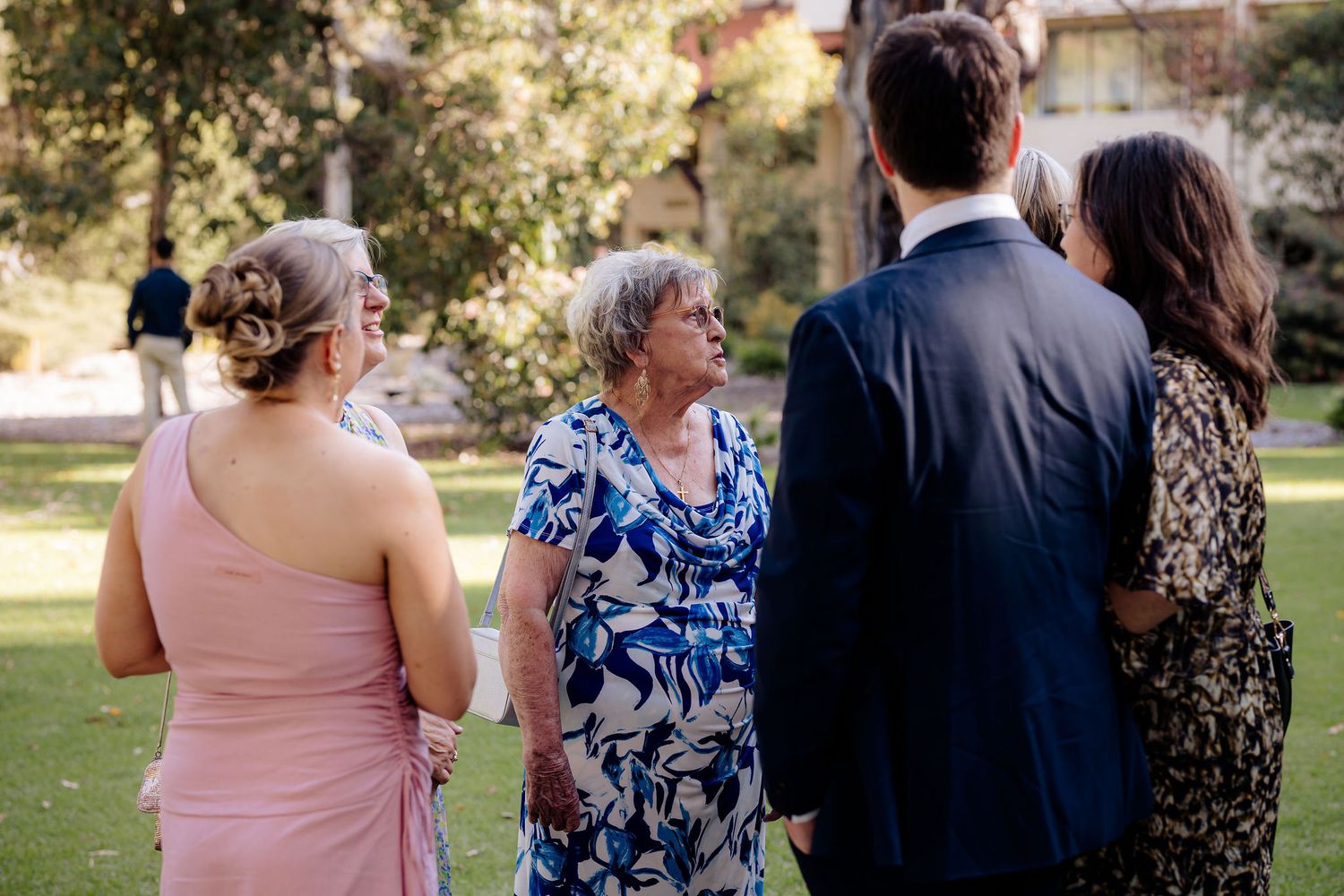 A small group gathers outdoors at a sunny event, with attendees wearing formal attire in pink, blue floral, and black.