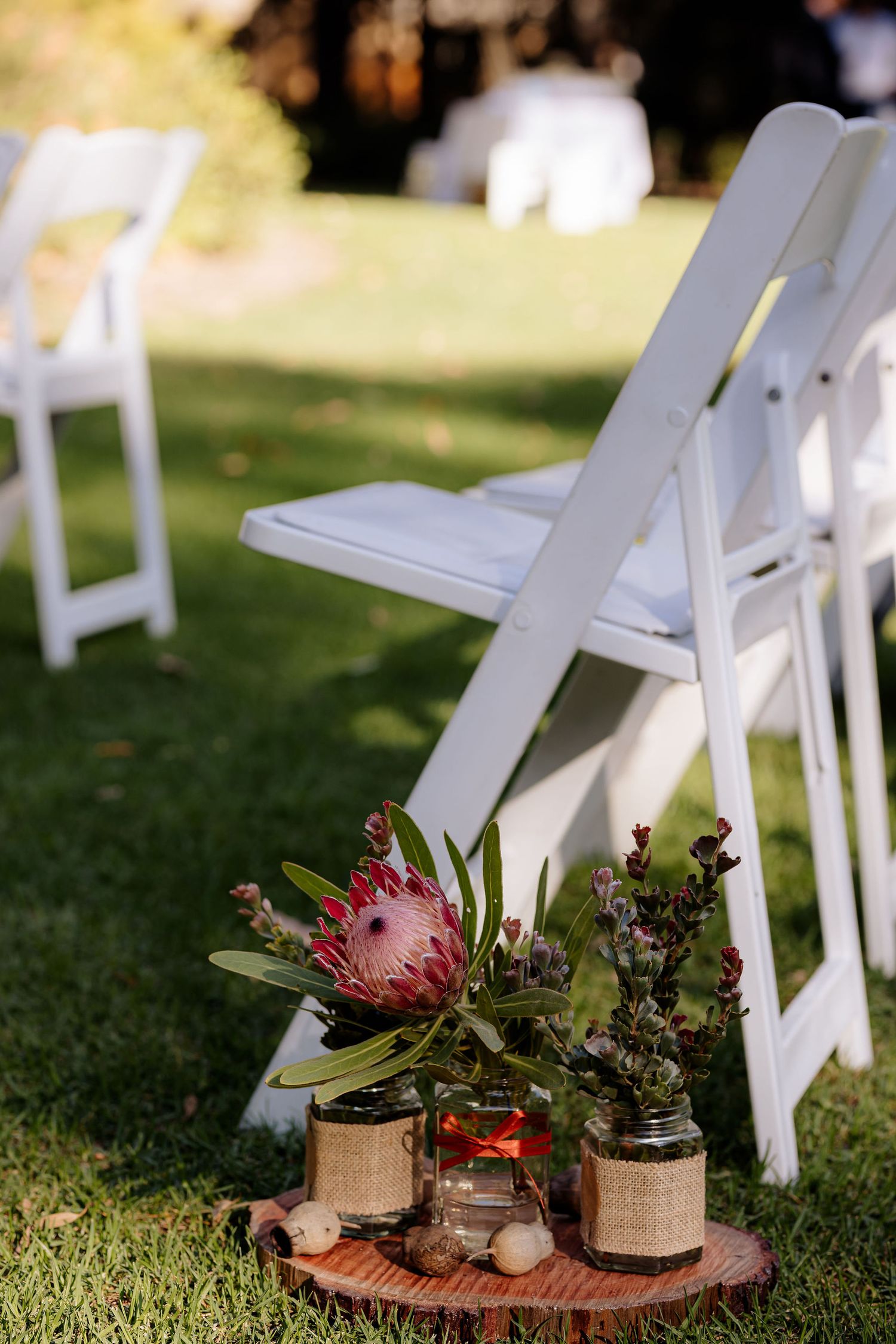White wooden folding chairs on grass lawn with rustic floral decor featuring protea flowers and burlap wrapped jars.