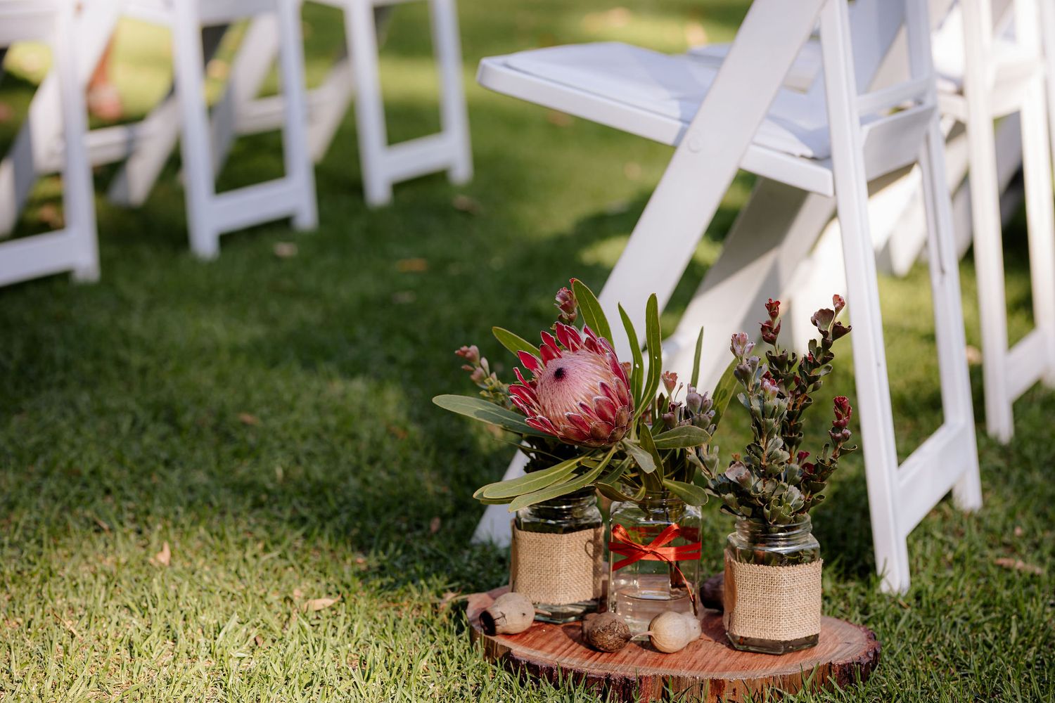 White ceremony chairs arranged on a lush grass lawn with potted flower arrangements adorning the aisle for an outdoor wedding.
