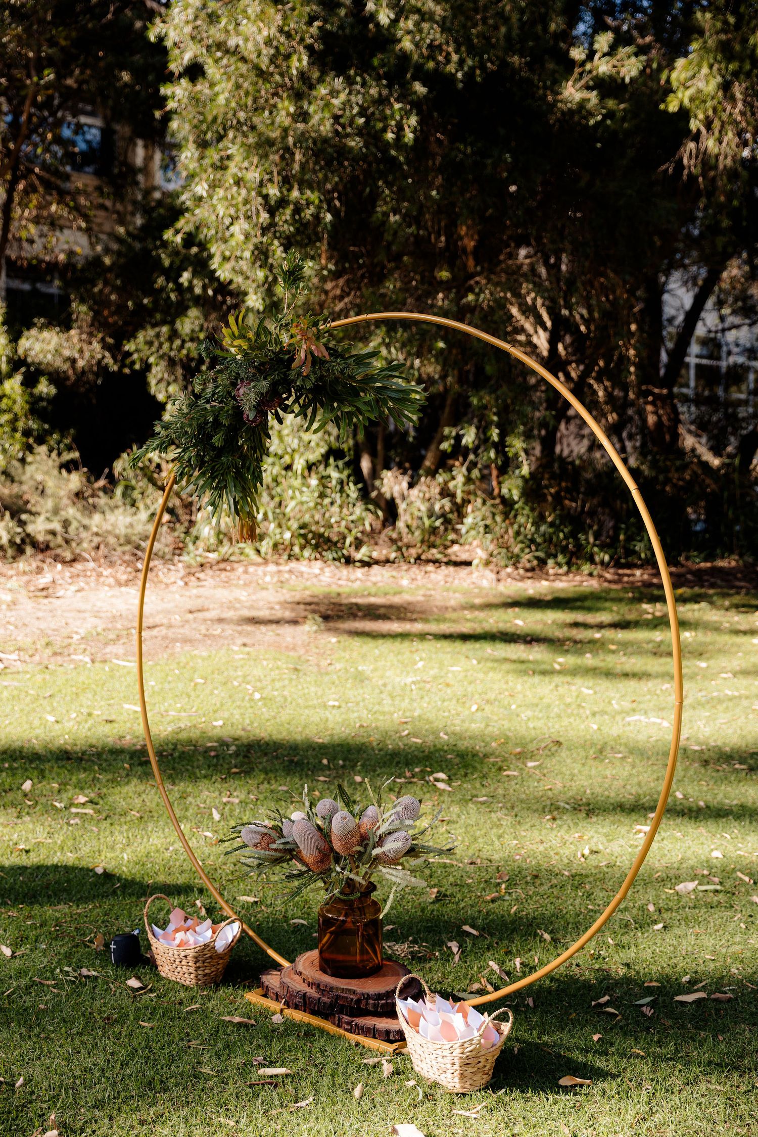 Gold circular wedding arch with floral arrangements and crystal vases on a sunny outdoor lawn.