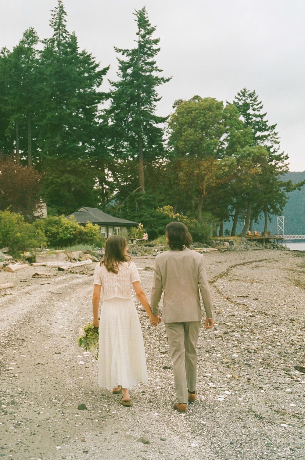 A couple strolls hand in hand along a rocky beach path with towering evergreens in the background.