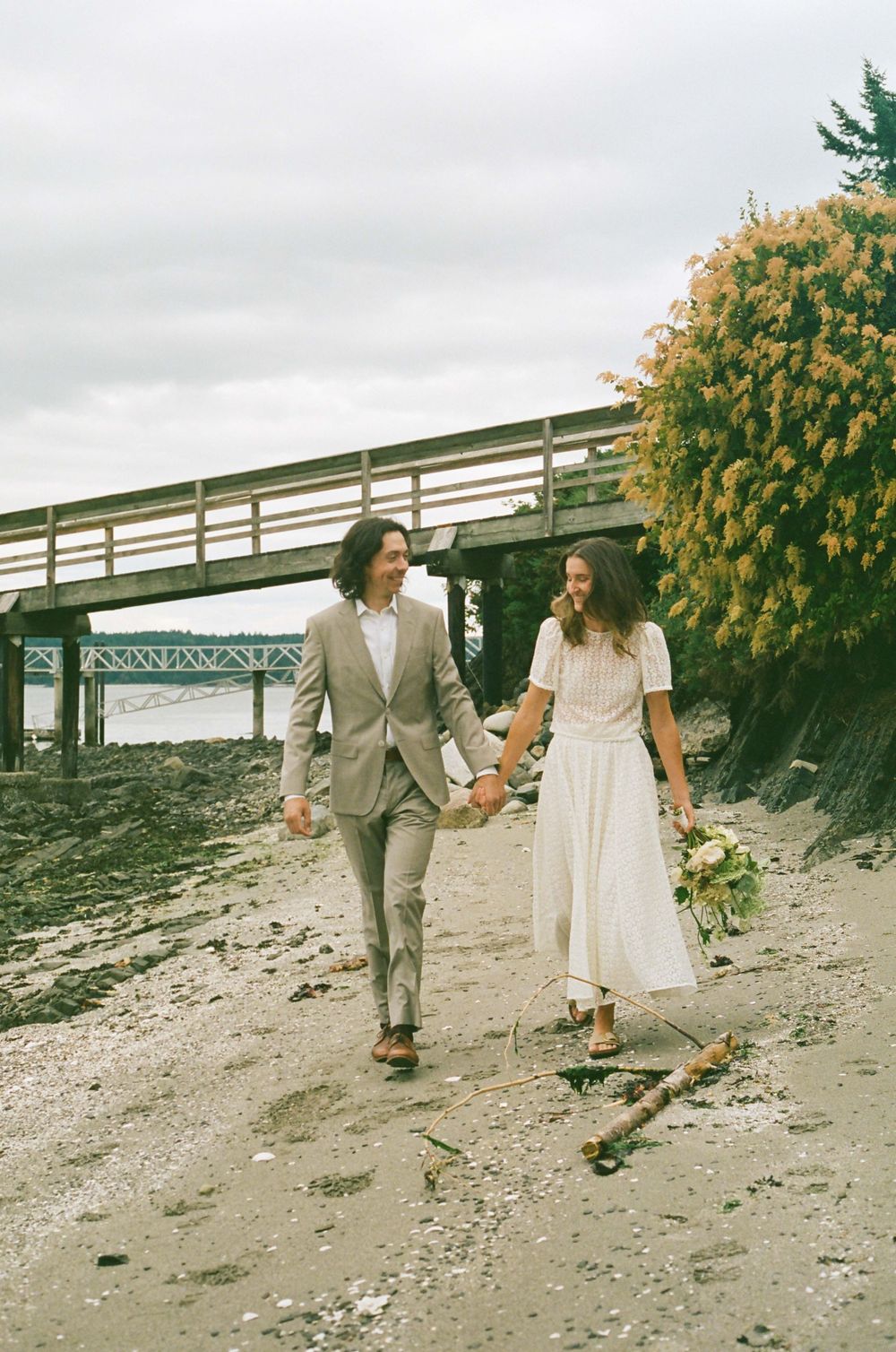 A couple walks beneath a wooden bridge on a rocky beach holding hands.