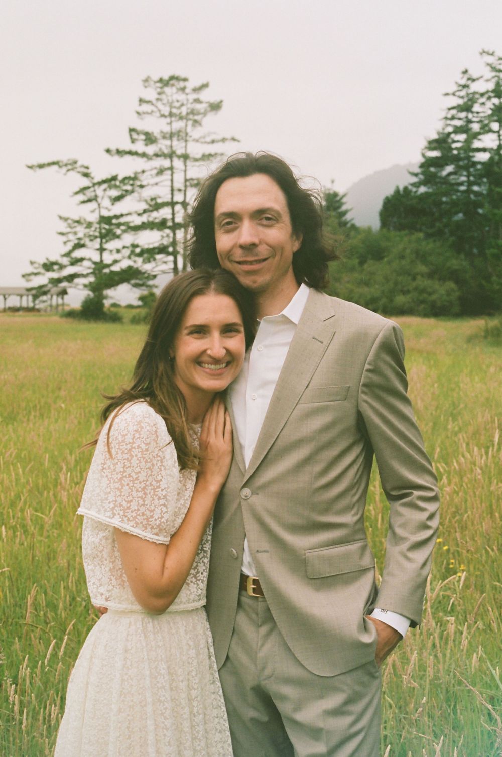 A couple poses together in a grassy field with trees, wearing white lace dress and beige suit.