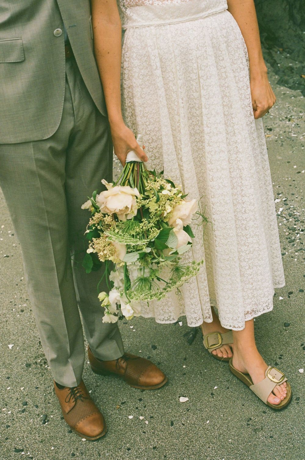 Close-up of white lace dress and simple bouquet with brown shoes on gravel ground.