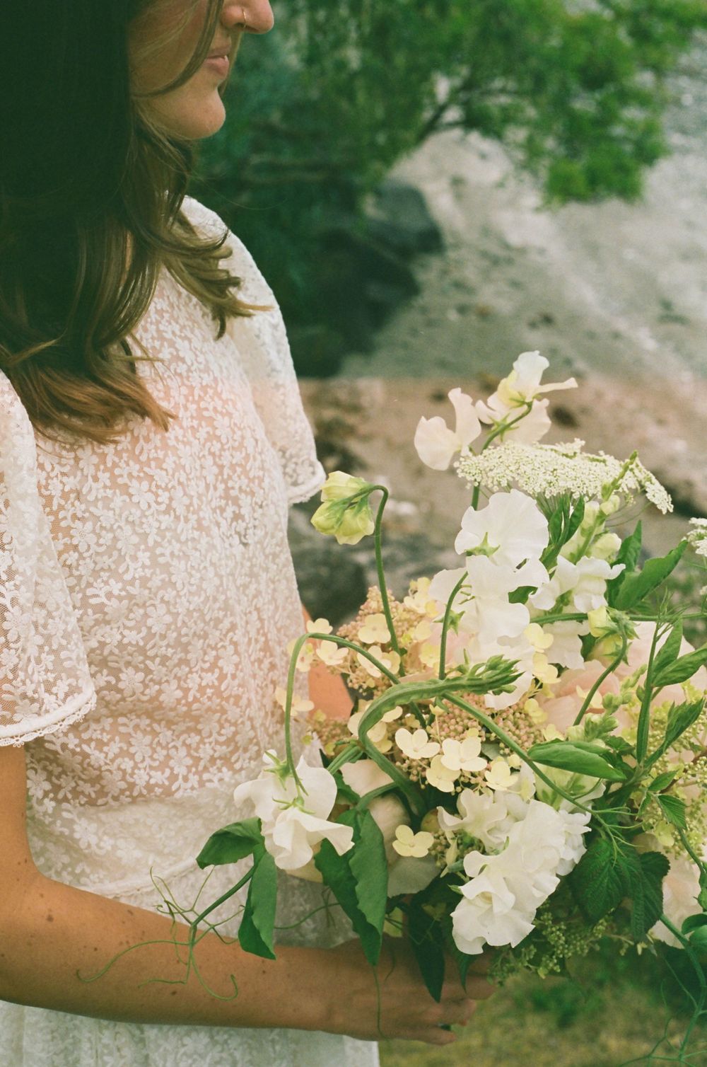 A delicate lace dress and romantic white floral bouquet create an ethereal bridal look against a natural backdrop.