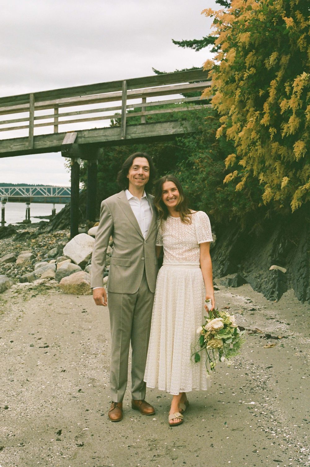Close-up of couple holding hands with a delicate white and green bouquet against rocky shoreline.