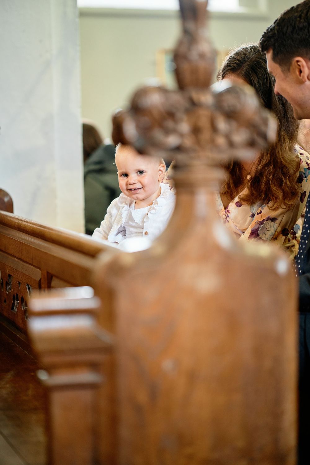 A smiling child in white baptismal clothing sits in a wooden church pew during a religious ceremony.
