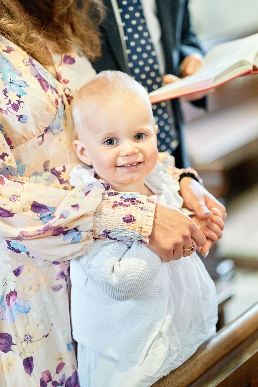 Small baby wearing a white dress smiles while sitting in caregiver's lap wearing floral print dress.