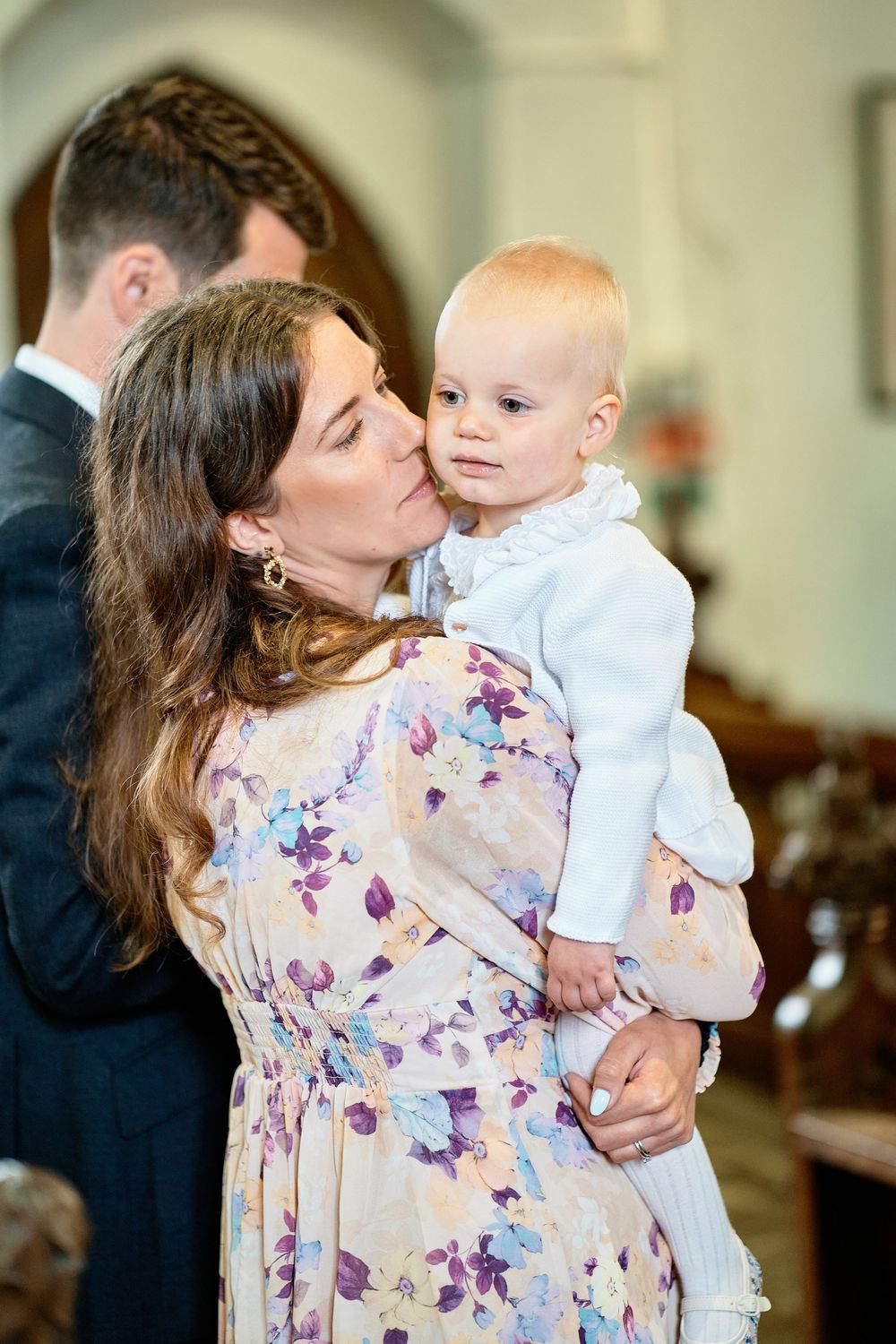 A woman in a floral dress lovingly holds and kisses a baby wearing white at an indoor gathering.