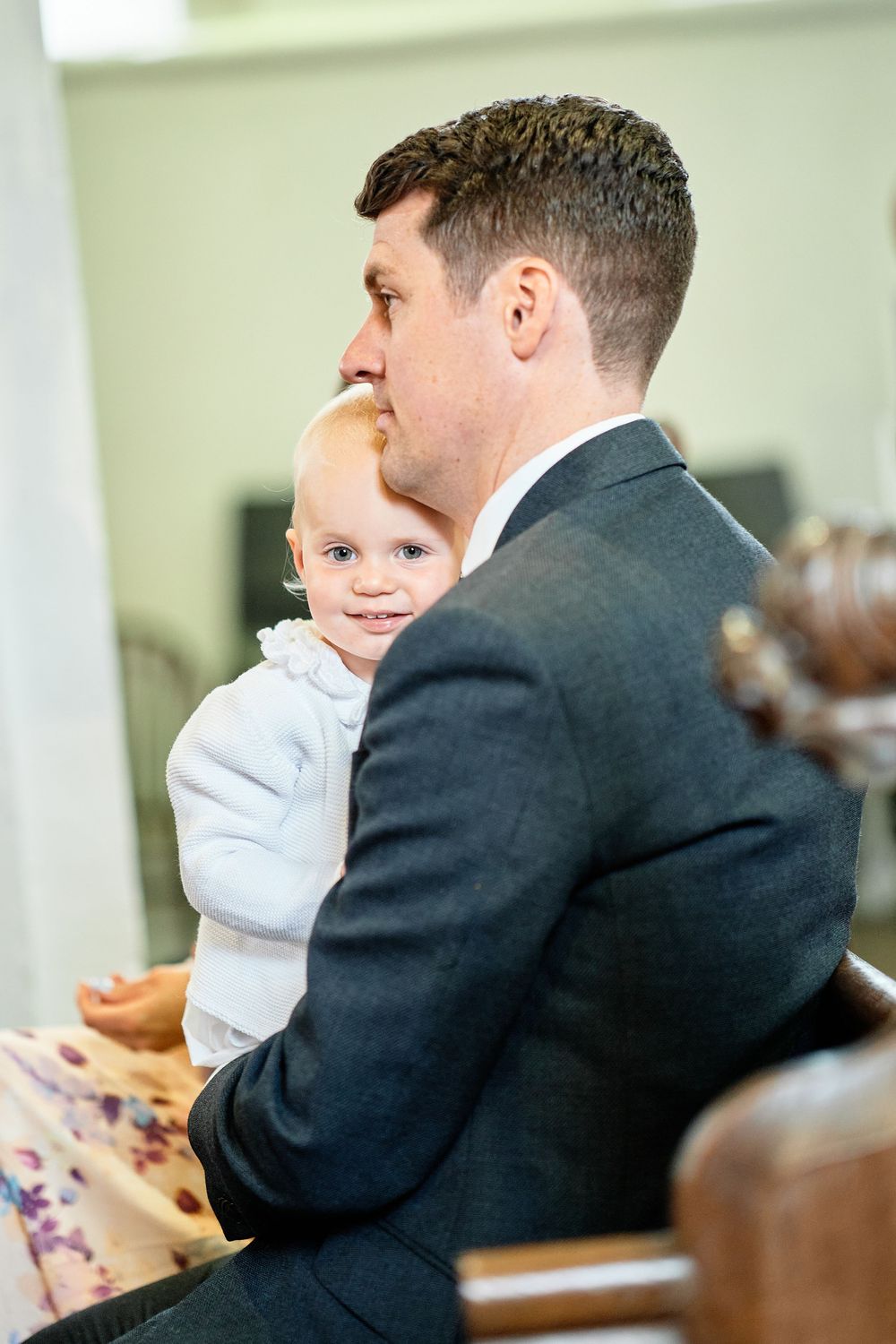 A parent in a dark suit holds a small child wearing white during what appears to be a formal event.