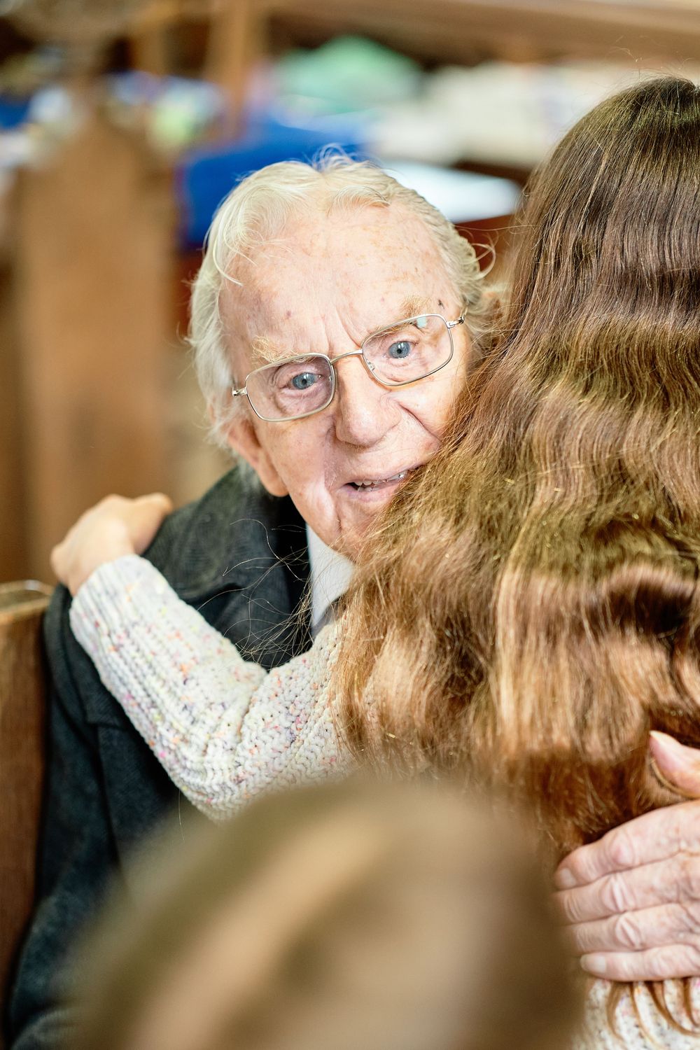 An elderly person wearing glasses and a sweater sharing a warm moment with someone in a blurry indoor setting.