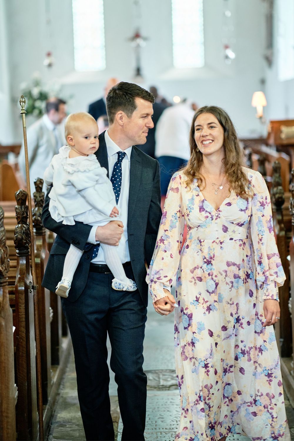 A couple walks down a church aisle with their baby, the mother wearing a floral dress and father in a navy suit.