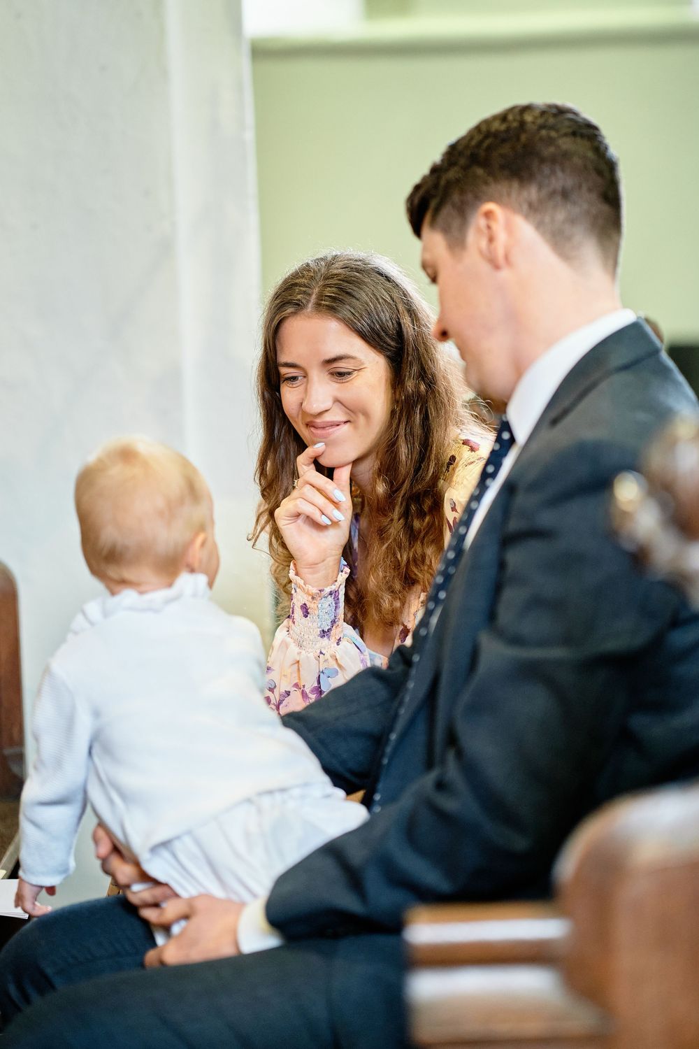 A couple shares a tender moment with a baby in formal attire at what appears to be a special event or ceremony.