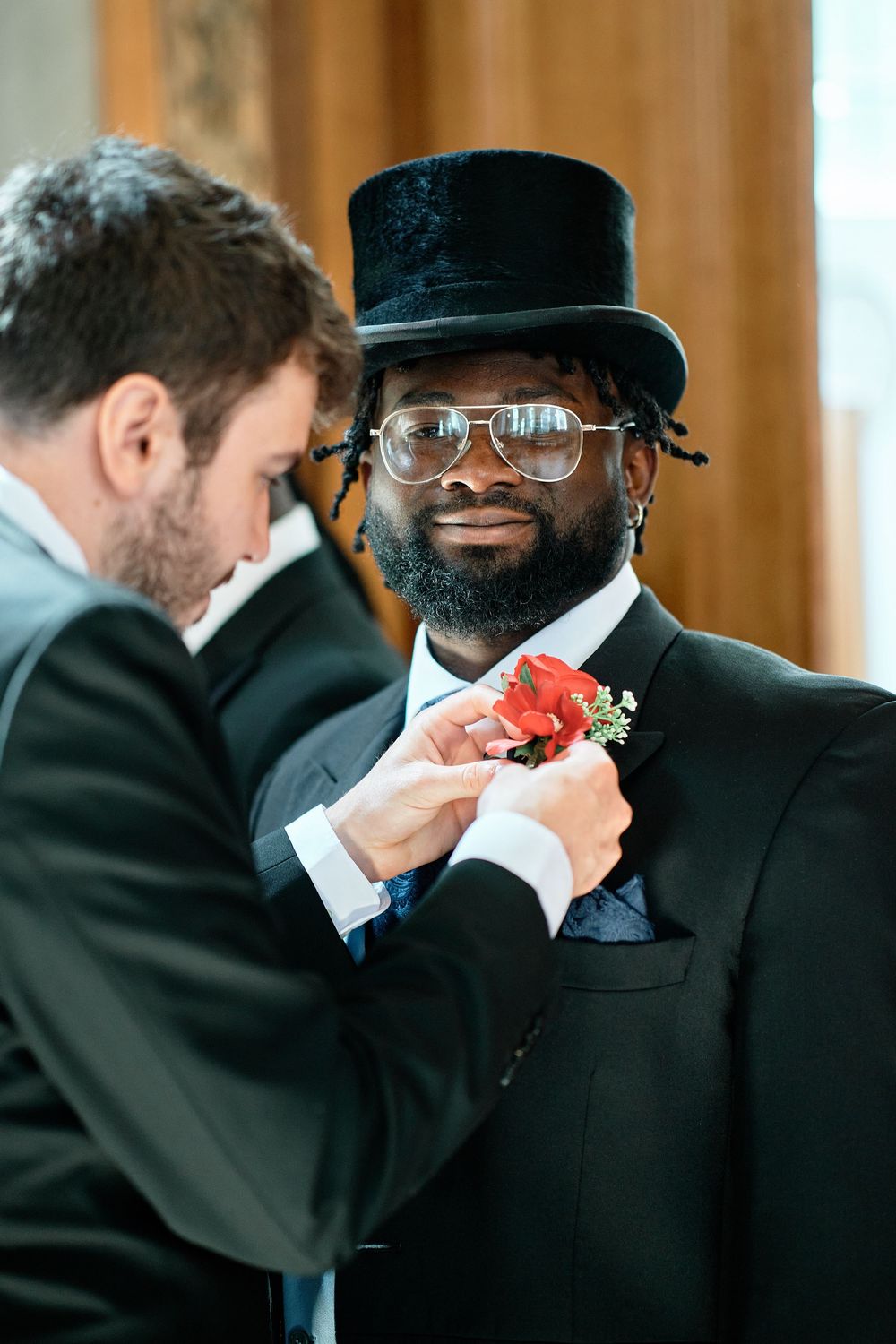 A groomsman wearing a top hat and suit receives help having a coral boutonniere pinned to his lapel.
