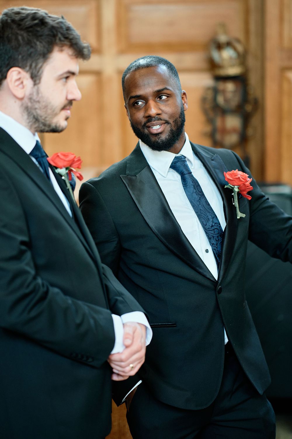 Two groomsmen in black tuxedos and red boutonnieres share a lighthearted moment during a wedding celebration.