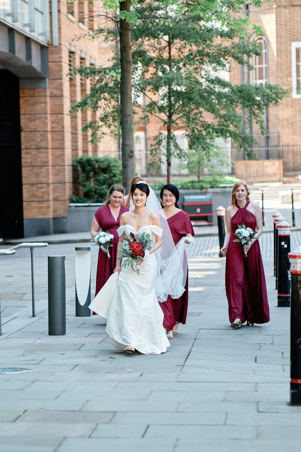 A sequence of wedding party photos shows playful moments along a brick building sidewalk with people in burgundy and white attire.
