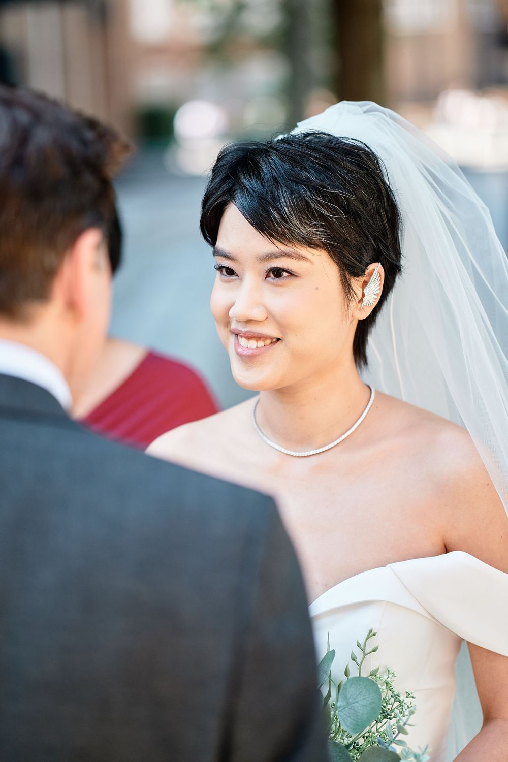 Beautiful bride in off-shoulder wedding dress and short hair wearing delicate necklace smiles during ceremony.