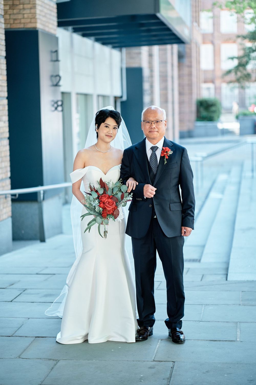 A bride in a white wedding gown walks down a covered outdoor walkway with someone by her side holding a red bouquet.