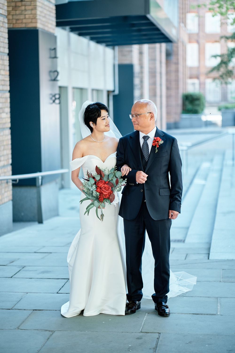 Two wedding attendees walk arm in arm on a city sidewalk, the bride in white holding a red bouquet of flowers.