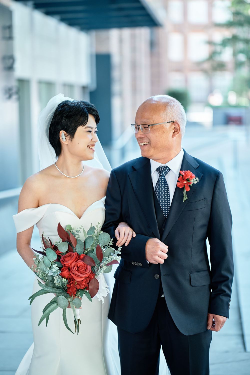 A bride in a white dress walks arm in arm with an older gentleman wearing a dark suit and red boutonniere.