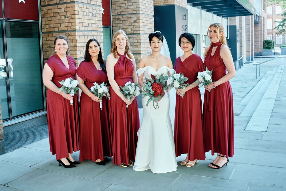 A bridal party poses outside on a sidewalk wearing matching burgundy gowns with the bride in traditional white.