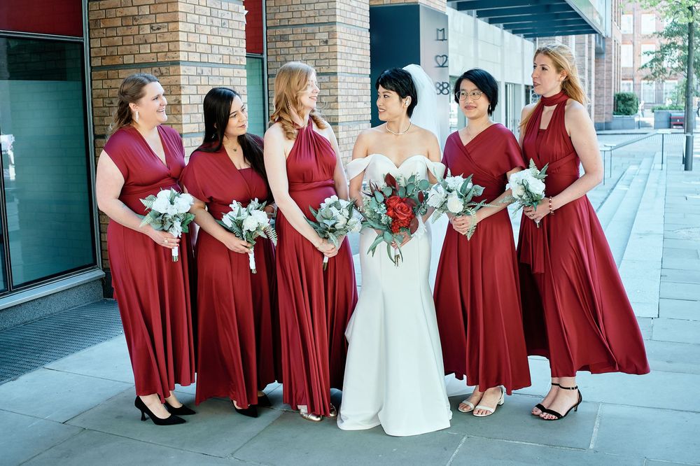 Wedding party in burgundy dresses pose together in an architectural setting with brick walls and columns.
