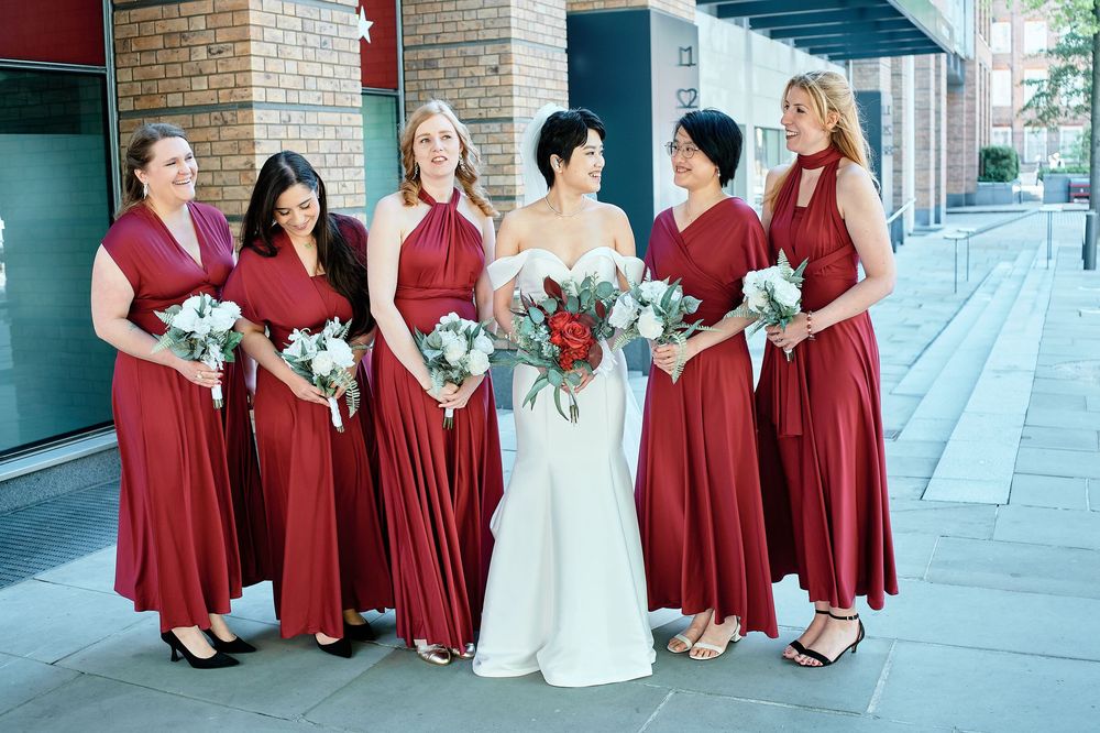 A wedding party in burgundy red bridesmaid dresses poses with the bride in a white wedding gown on a city sidewalk.