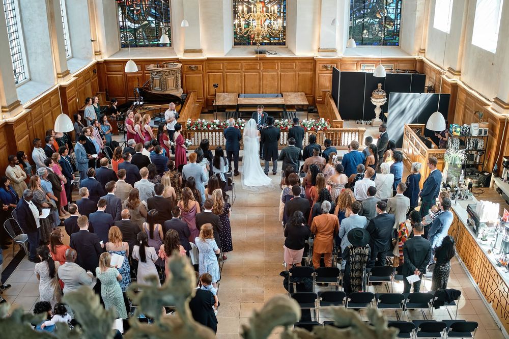 A wedding ceremony in progress inside a grand wood-paneled hall with guests standing and watching the proceedings.