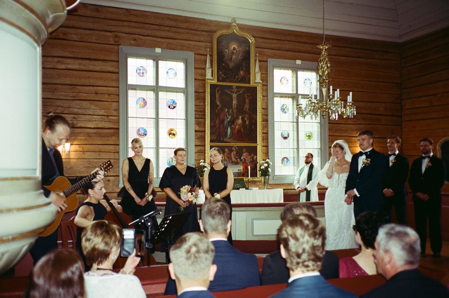 Wedding ceremony attendees gather in a rustic wooden-paneled room with windows and elegant lighting fixtures.