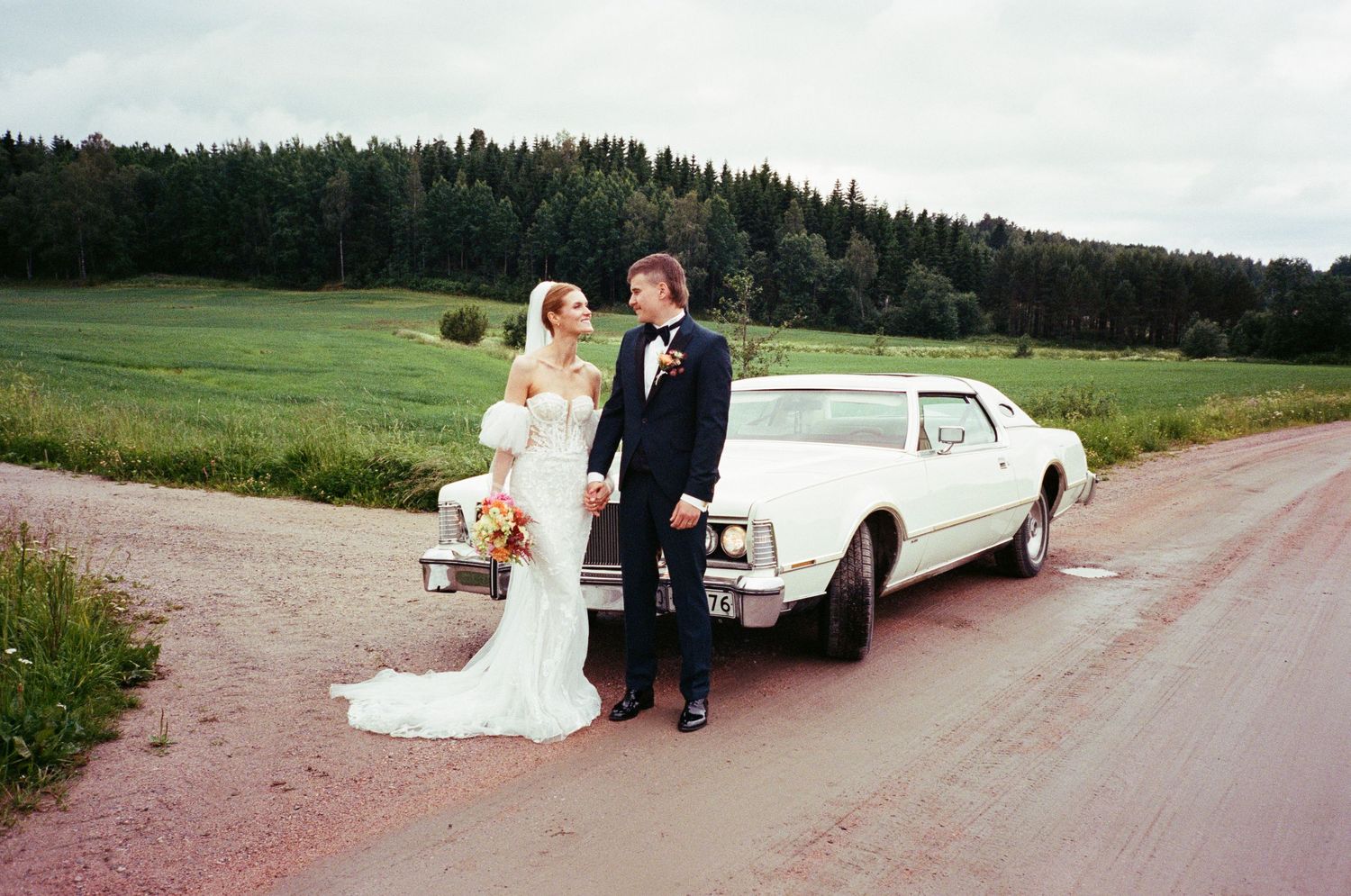 A panoramic view of wedding moments near a classic white car in a rustic countryside setting on a cloudy day.