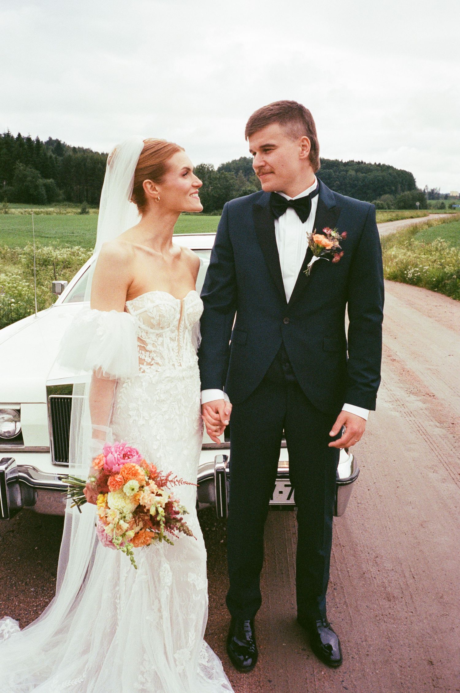 A newlywed couple walks along a rustic country road surrounded by green fields and trees in their formal attire.