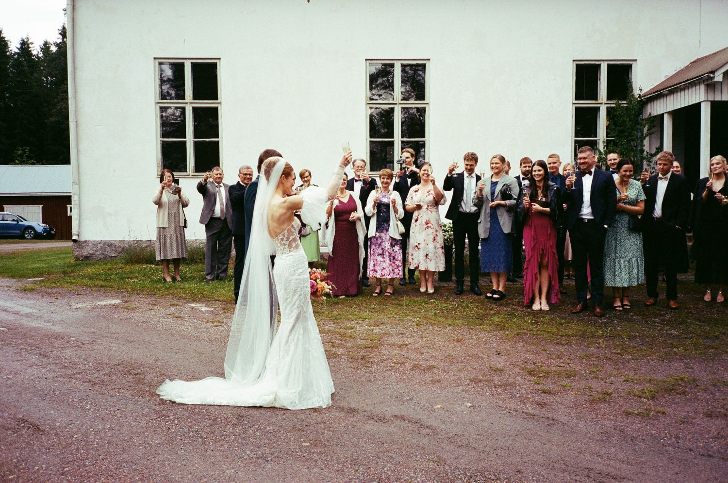 Wedding party and guests gather outside a white building as couple celebrates during an outdoor celebration.