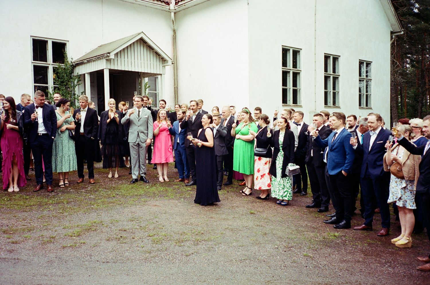 Large group of wedding guests in formal attire gathered outside a white church building for a celebration.