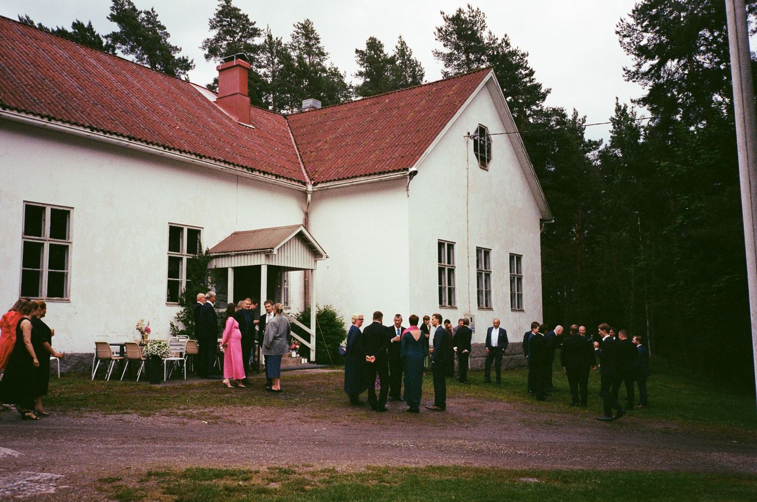 A group of people gathered outside a white building with a red roof during an event in a wooded area.
