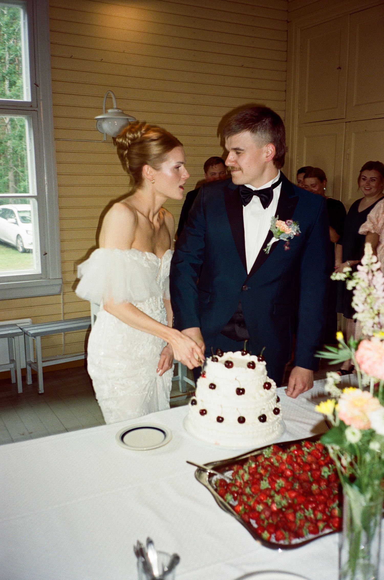 Tiered white wedding cake with chocolate decorations sits on table alongside platter of fresh red strawberries at reception.