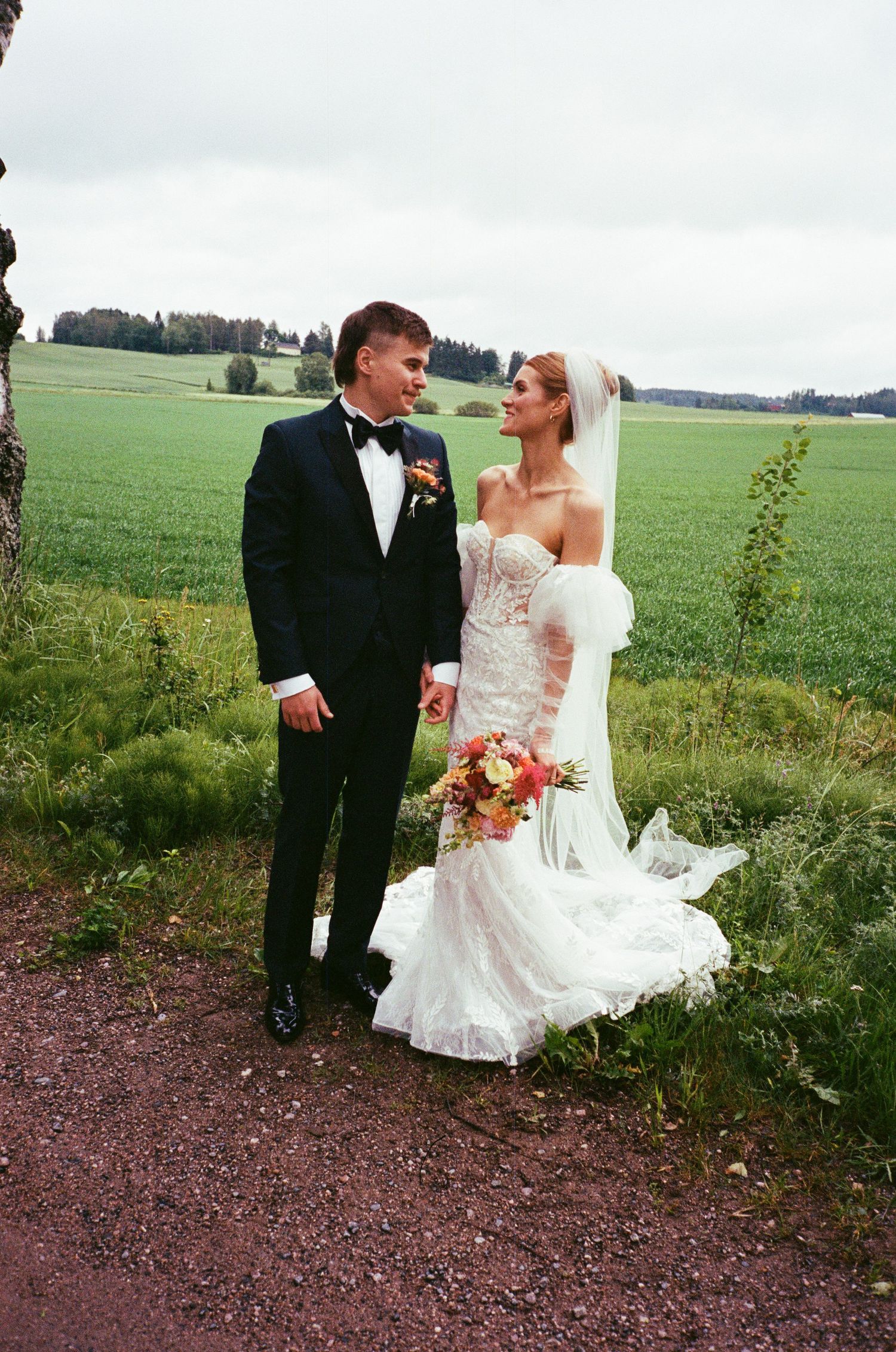 Couple in wedding attire share a romantic moment while posing on a dirt path next to green farm fields.