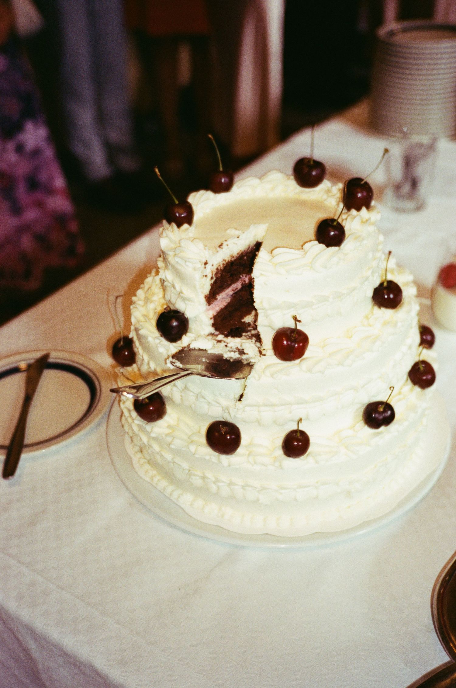 A white frosted cake decorated with chocolate shavings and dark cherries sits on a plate at what appears to be a bakery.