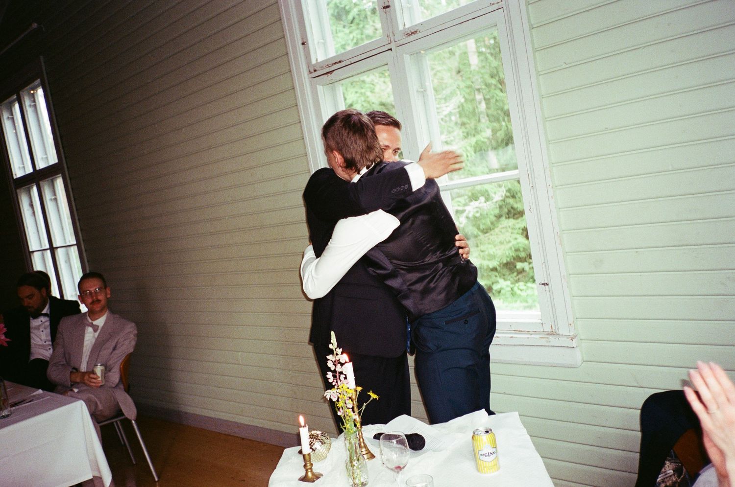 Two people embrace in an emotional moment during a casual indoor celebration with windows and table settings visible.
