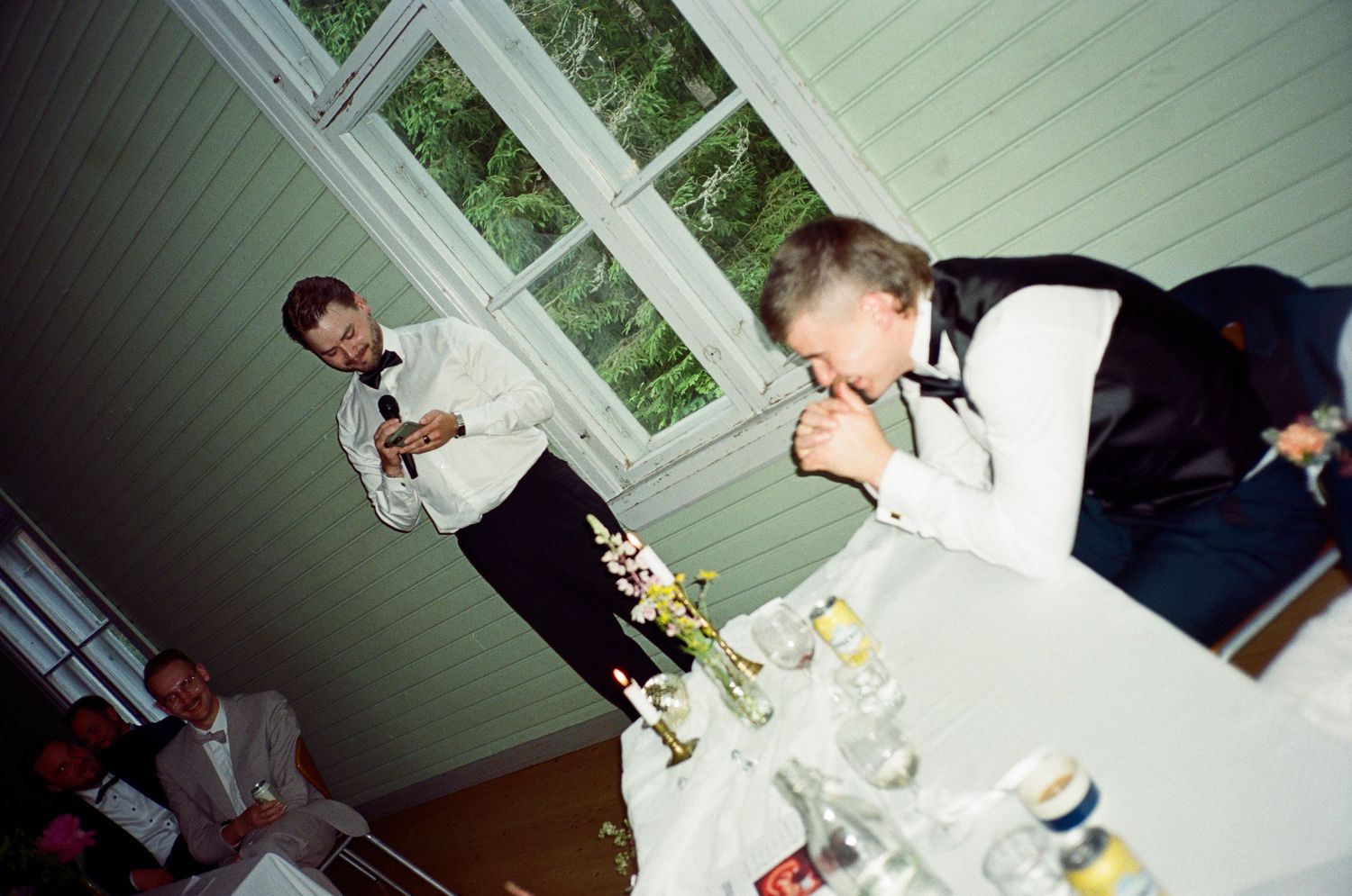 Two people partying indoors with beverages at a casual gathering near windows with greenery visible outside.