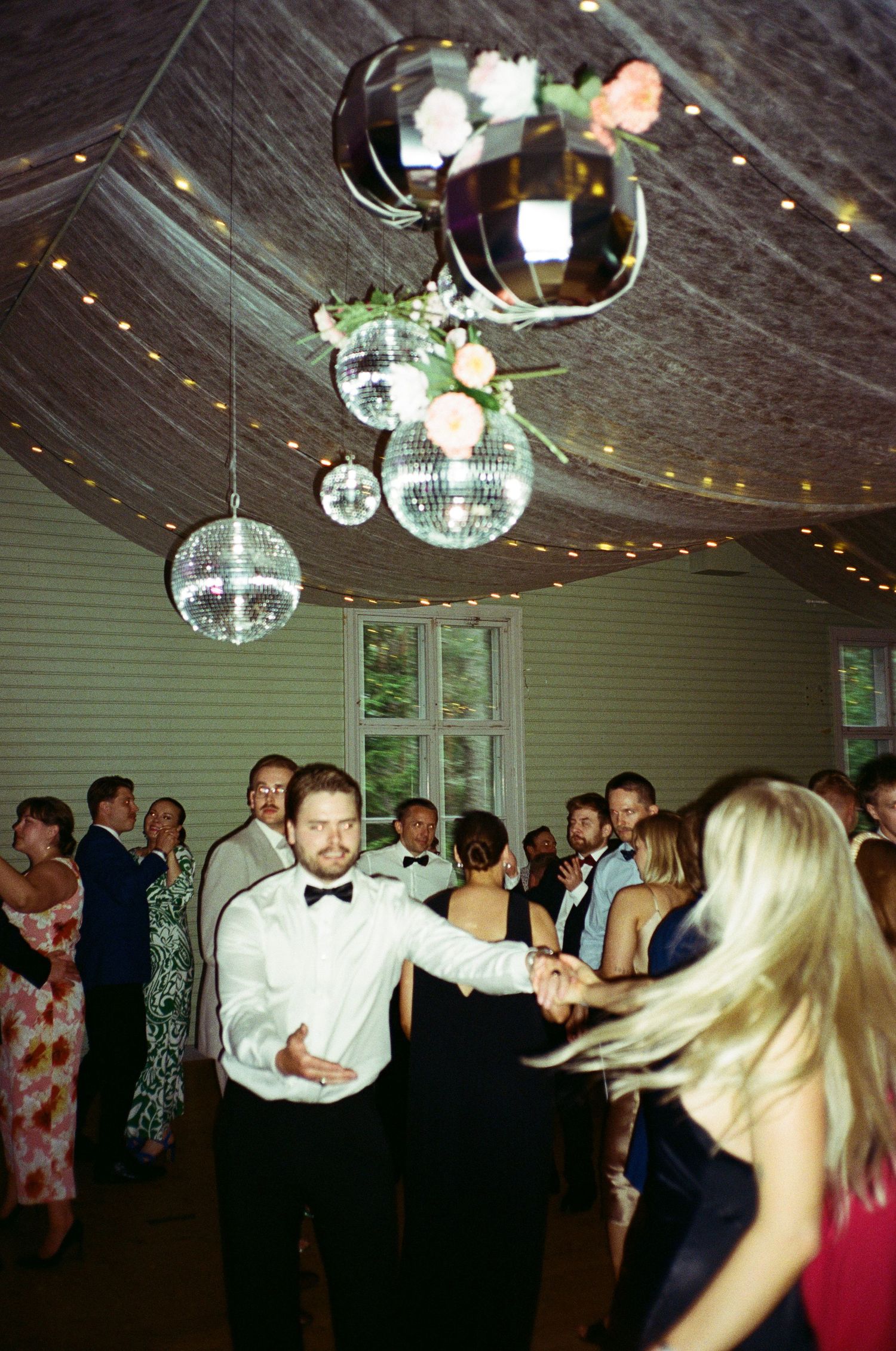 Guests dancing at a wedding reception under disco ball lighting and string lights in a rustic indoor venue.