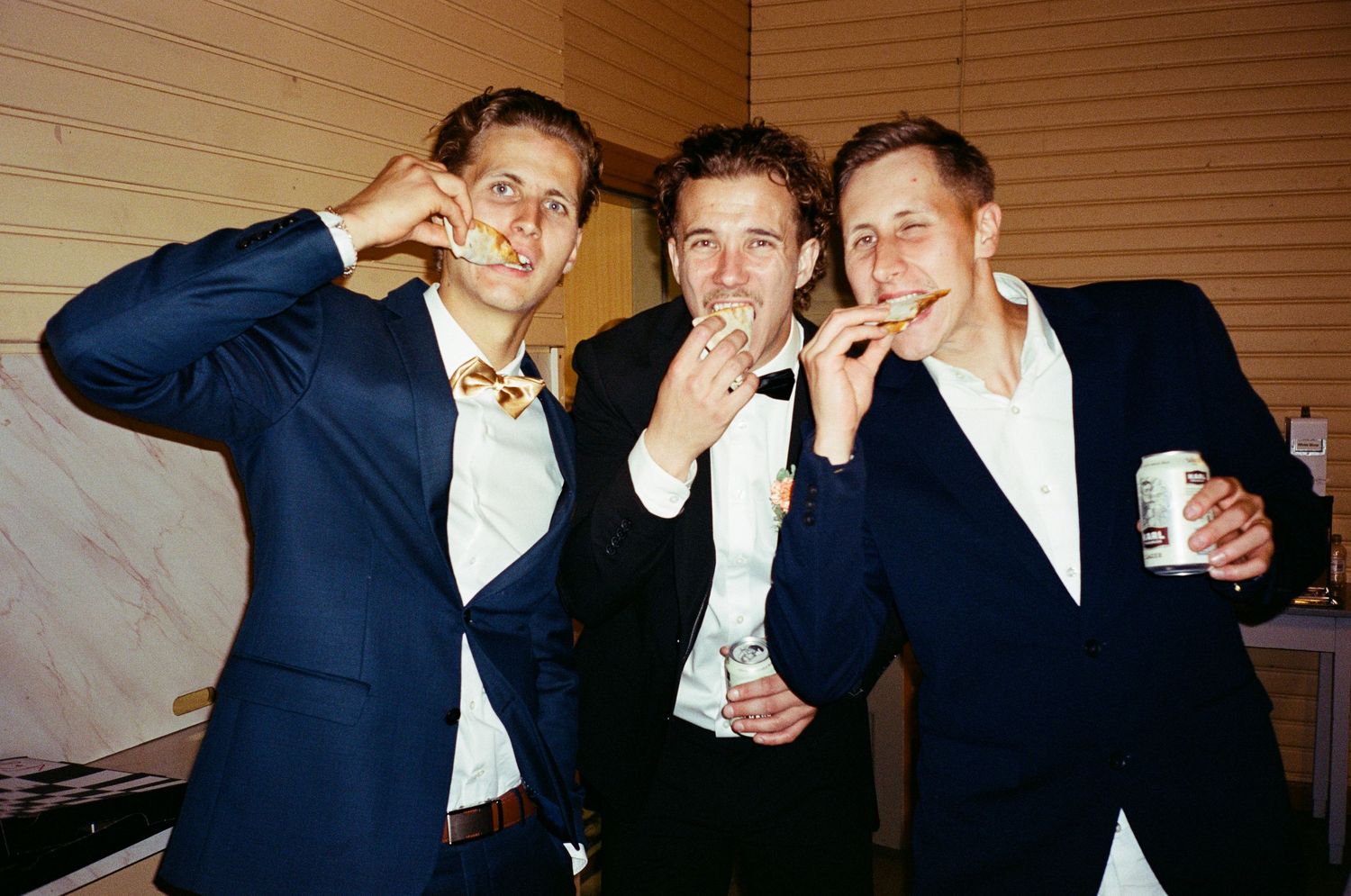 Three suited party-goers make silly faces and gestures while holding drinks at an indoor celebration.