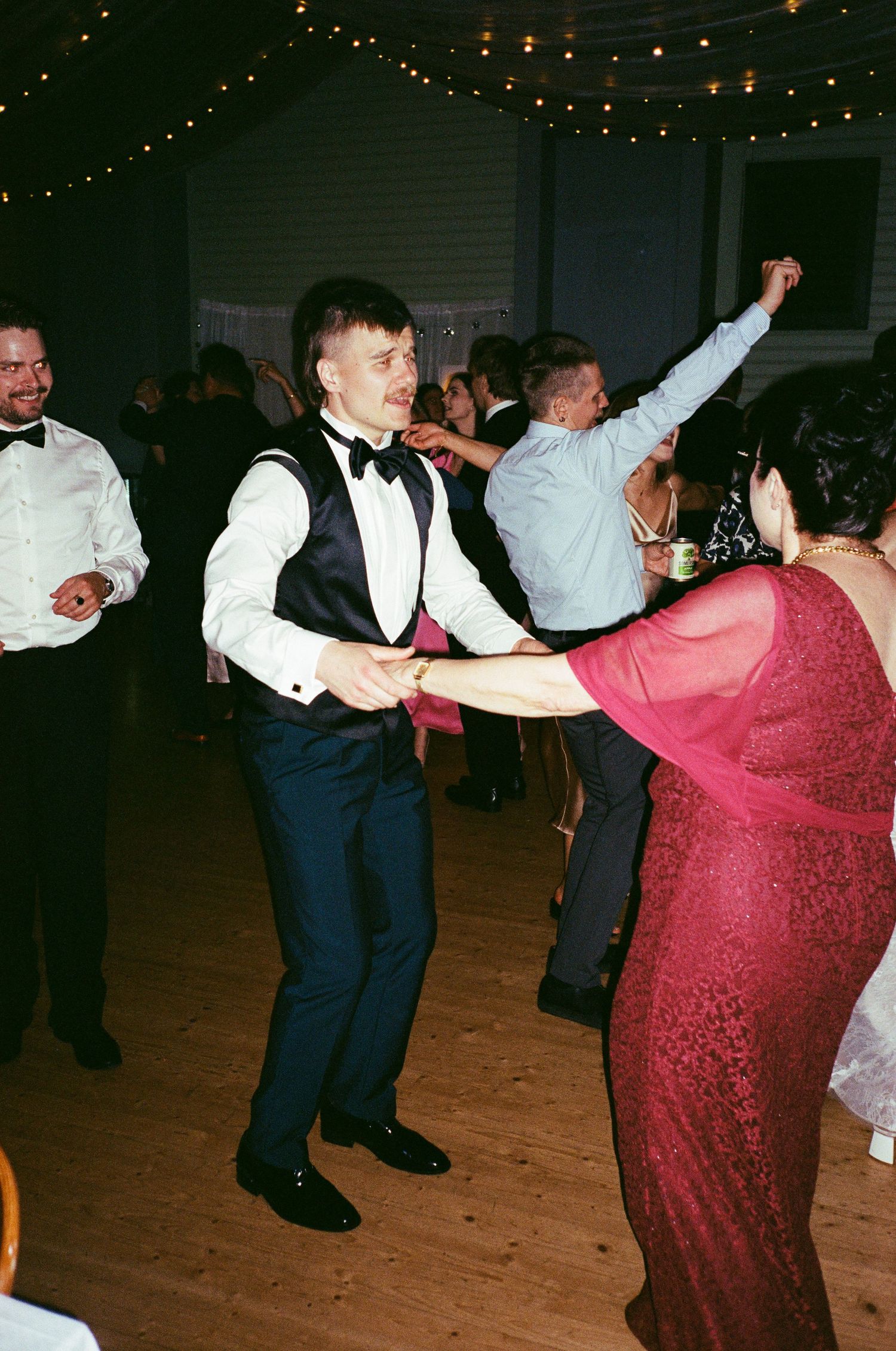 People dancing at a formal event with string lights overhead, featuring a person in a bow tie and vest dancing with someone in red.